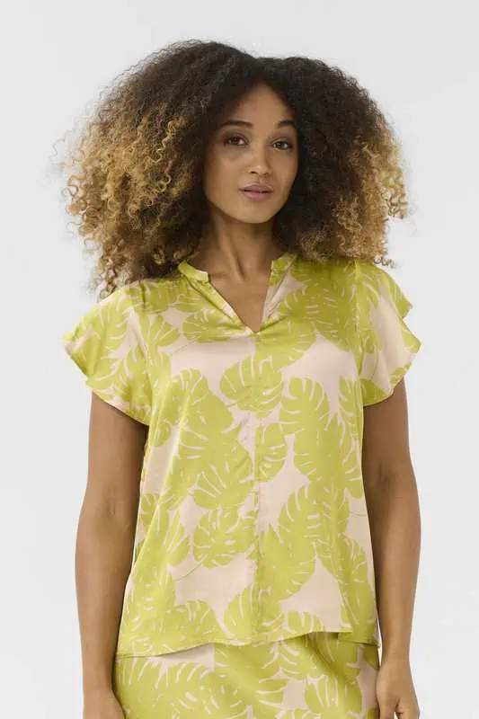 Kvinna bär en somrig blus med limegrönt monstera-mönster, kort fjärilsärm och V-ringning, stylad med matchande underdel.