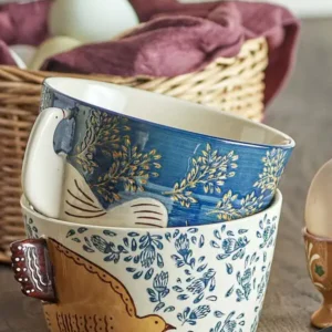 Två staplade Hezha-keramikskålar från Bloomingville – en blå med lövmönster och en beige med fågelmotiv, placerade på ett träbord med korg i bakgrunden.