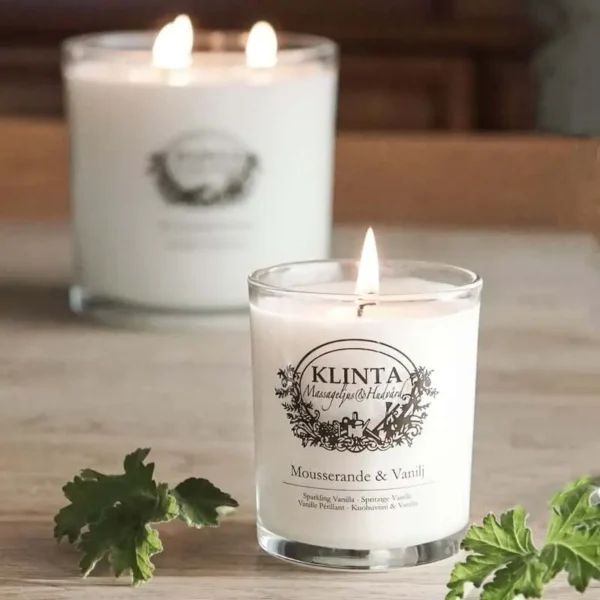 Tänt Klinta doft- och massageljus Mousserande & Vanilj på trägolv med gröna blad och större ljus i bakgrunden.