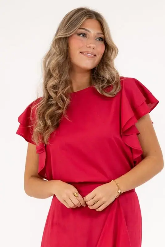 Kvinna i röd blus med volangärmar och rund hals från Capri Collection, poserar leende med händerna i midjan.
