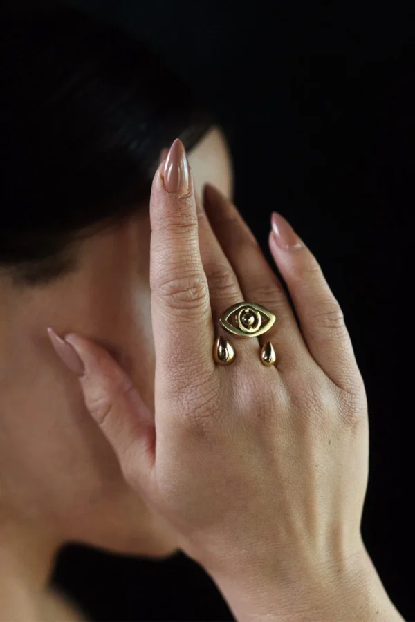Voyance ring i guldpläterat stål med öga och tårar bärs på hand med ljus manikyr, fotograferad mot svart bakgrund.