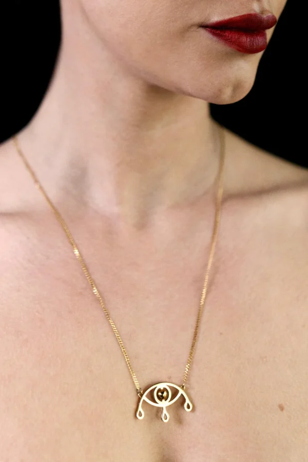 Voyance halsband i guld med öga och tre tårar bärs av modell med bar hud och rött läppstift, fotograferad i närbild mot svart bakgrund.