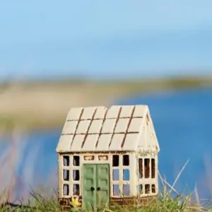 Litet stengodshus med glasruteformade fönster och grön dörr, stående i gräs vid vattnet.