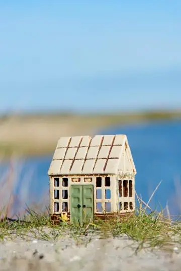 Litet stengodshus med glasruteformade fönster och grön dörr, stående i gräs vid vattnet.