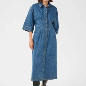 Kvinna i lång medelblå denimklänning med trekvartsärmar och knappar framtill, stylad med svarta stövlar.