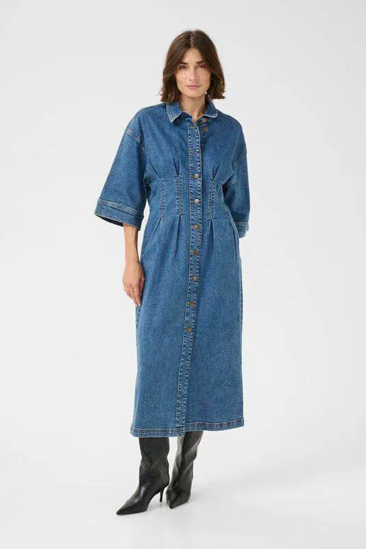 Kvinna i lång medelblå denimklänning med trekvartsärmar och knappar framtill, stylad med svarta stövlar.