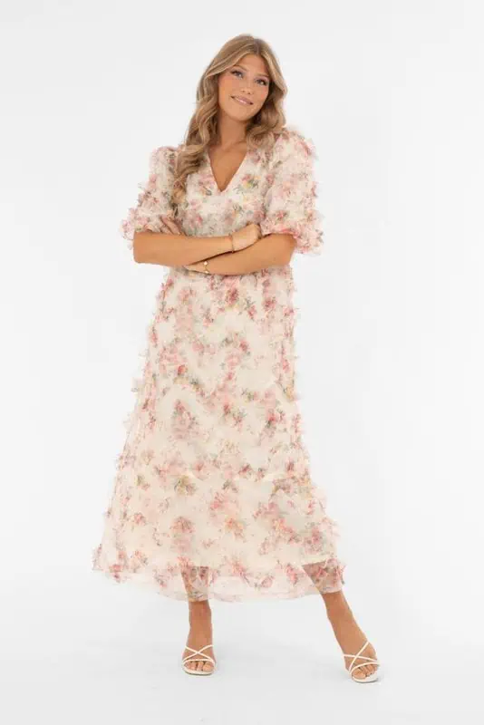 Modell bär lång blommig klänning i buttercream och rosa från Capri Collection med v-ringning, puffärmar och luftig kjol.