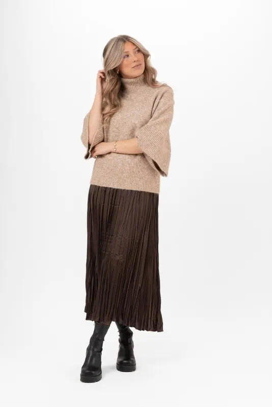 Modell i lång mörk plisserad satinkjol från Capri Collection, brun stickad polotröja och svarta boots mot vit bakgrund.