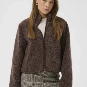 Modell i brunmelerad CUcatina Teddy Jacket från Culture med mjuk teddystruktur, hög krage, dragkedja och långa ärmar, matchad med rutig kjol.